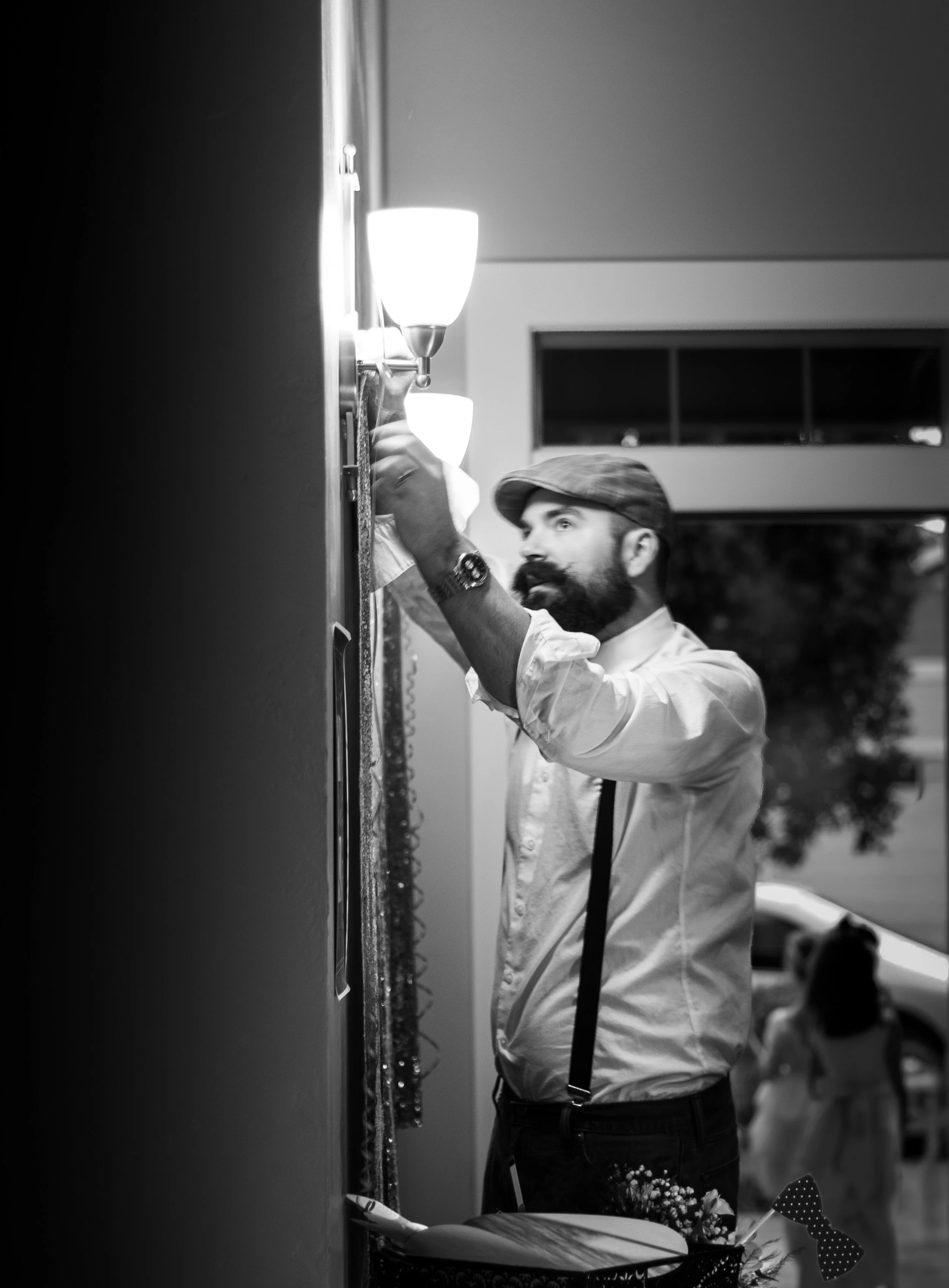 A man with a beard wearing a flat cap and suspenders decorates a wall with lights at night.