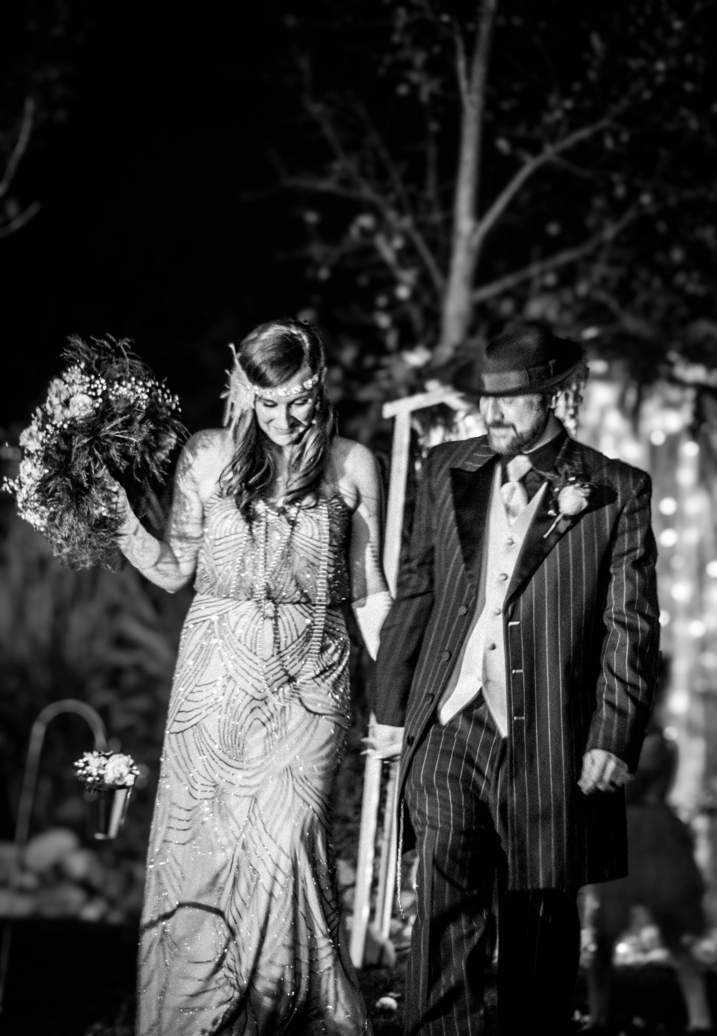 Black and white photo of a bride and groom holding hands during their wedding ceremony at night outdoors, with decorative lighting and trees in the background.