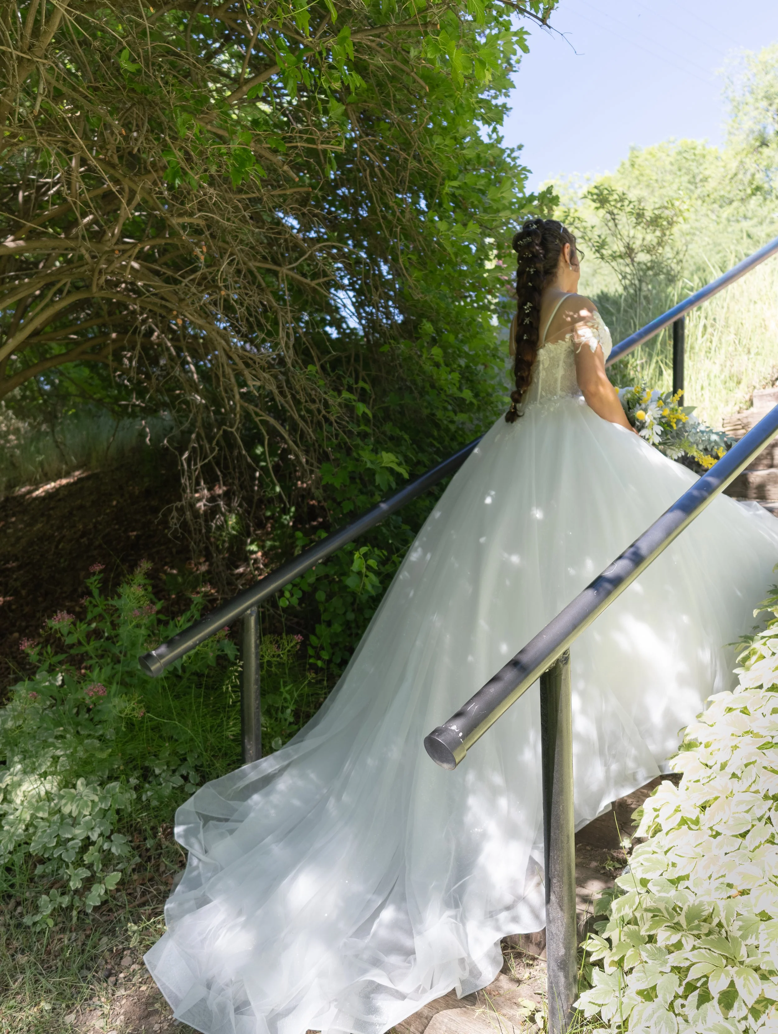 A woman in a white wedding dress with lace details on the sleeves, sitting on outdoor stairs, holding a bouquet, with her hair styled in a long braid decorated with small white flowers, surrounded by green foliage and white flowers.