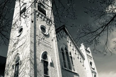 A tall, historic church with a clock tower and arched windows in black and white, surrounded by tree branches.