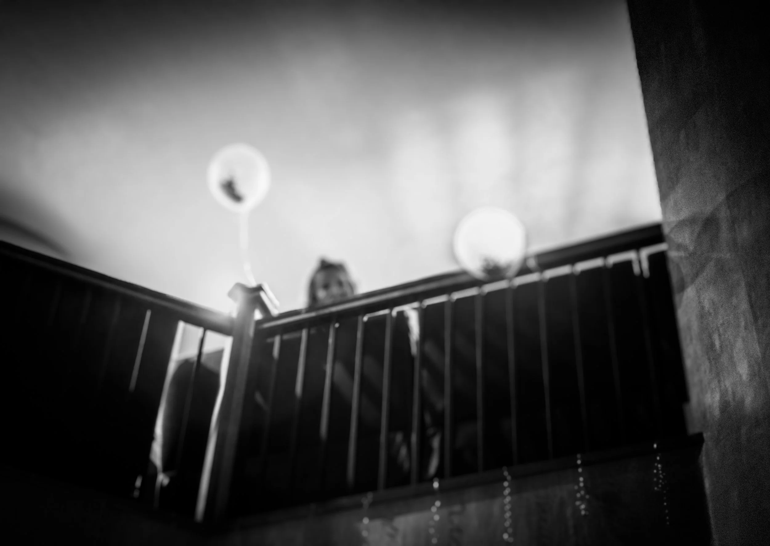 Black and white photo of a person leaning on a balcony railing, taken from below the person, with balloons in the background.