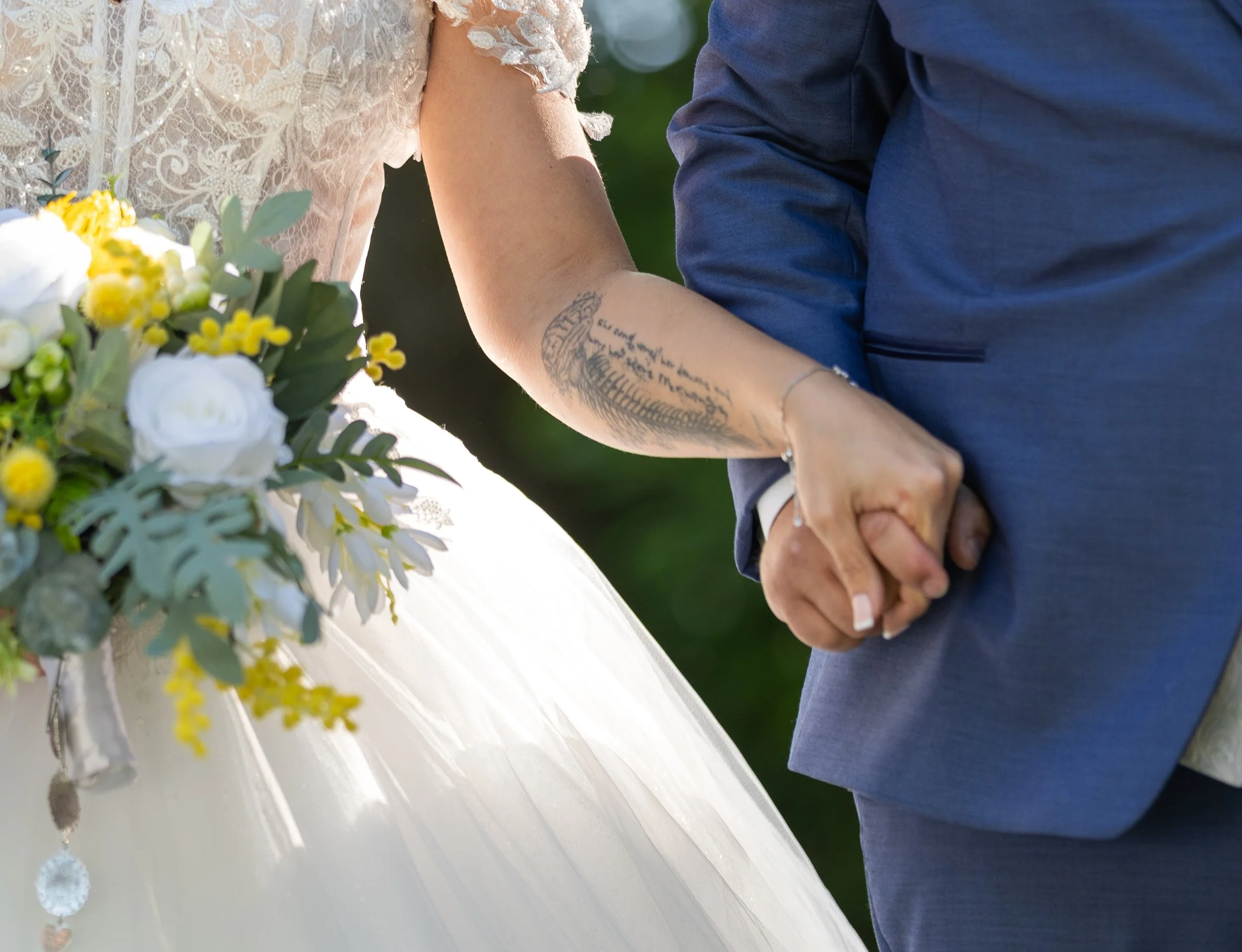 Close-up of a couple holding hands during a wedding ceremony, with the bride in a lace dress and the groom in a blue suit, and part of a bouquet of white and yellow flowers in the foreground.