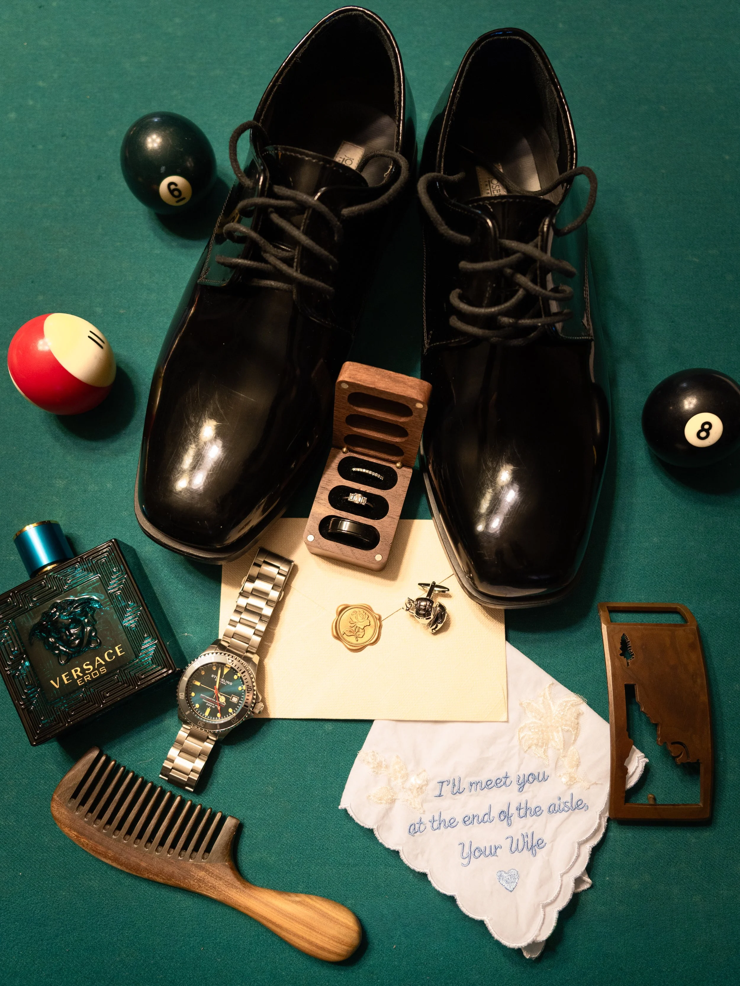 Black shiny men's dress shoes on a green surface, alongside billiard balls, a wristwatch, a hair comb, a Versace perfume box, a decorative wooden block, a handwritten note, and various small objects.