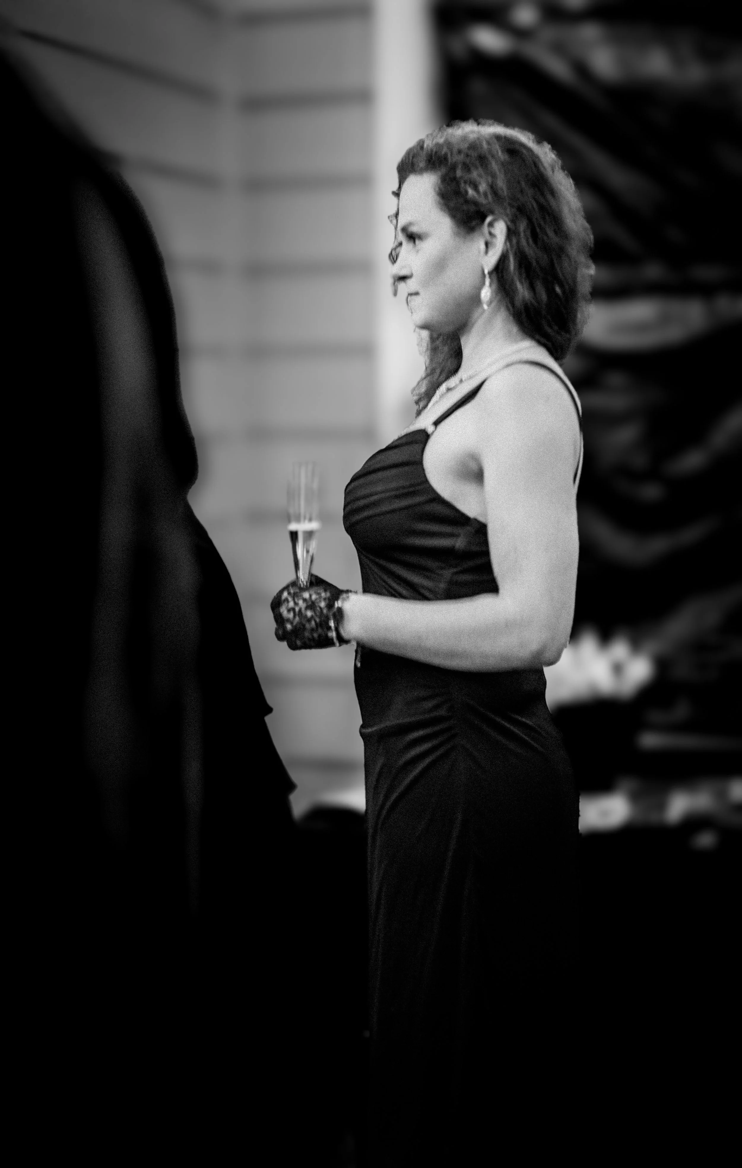 A woman in a black evening gown holding a glass of champagne at a formal event, with a focused expression.