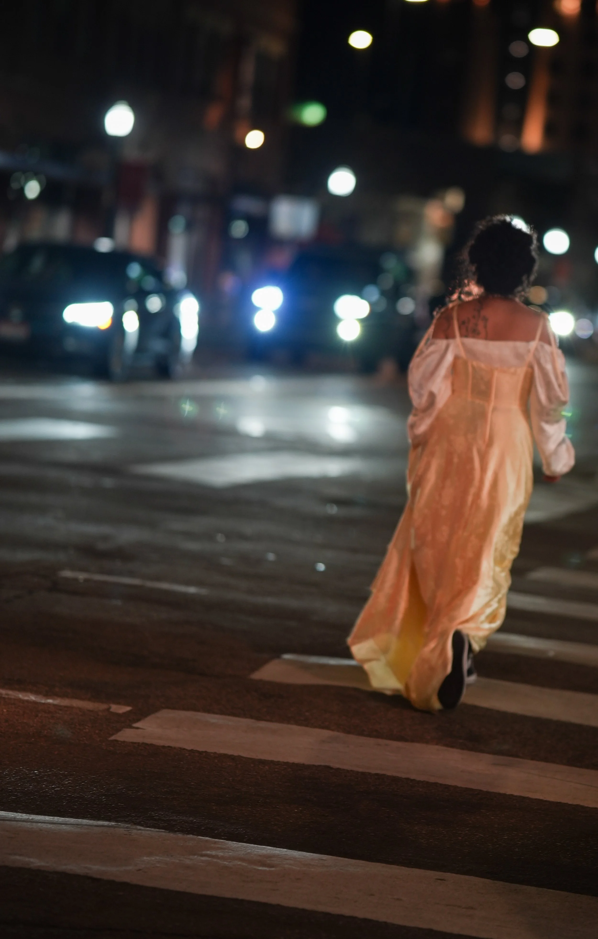 A woman with curly hair, wearing a yellow dress, walking across a city street at night on a crosswalk with blurred cars and bright lights in the background.