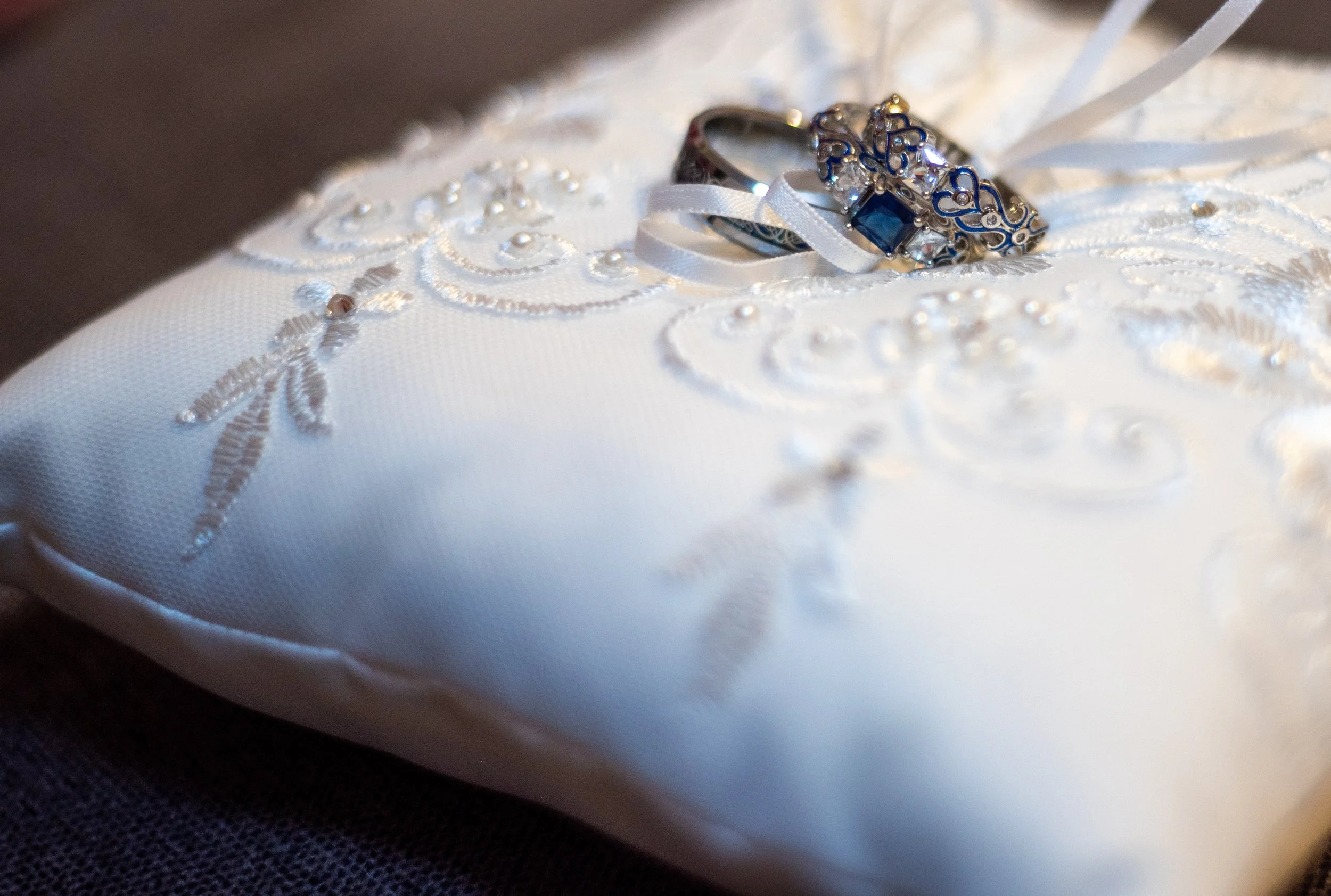 Close-up of a white satin pillow with wedding rings resting on top, embroidered with white thread and small pearls, on a dark surface.