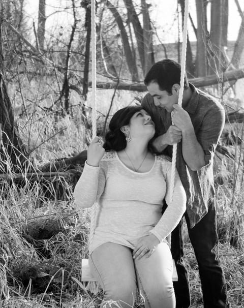 A couple sitting on a swing in a wooded area, smiling and looking at each other.