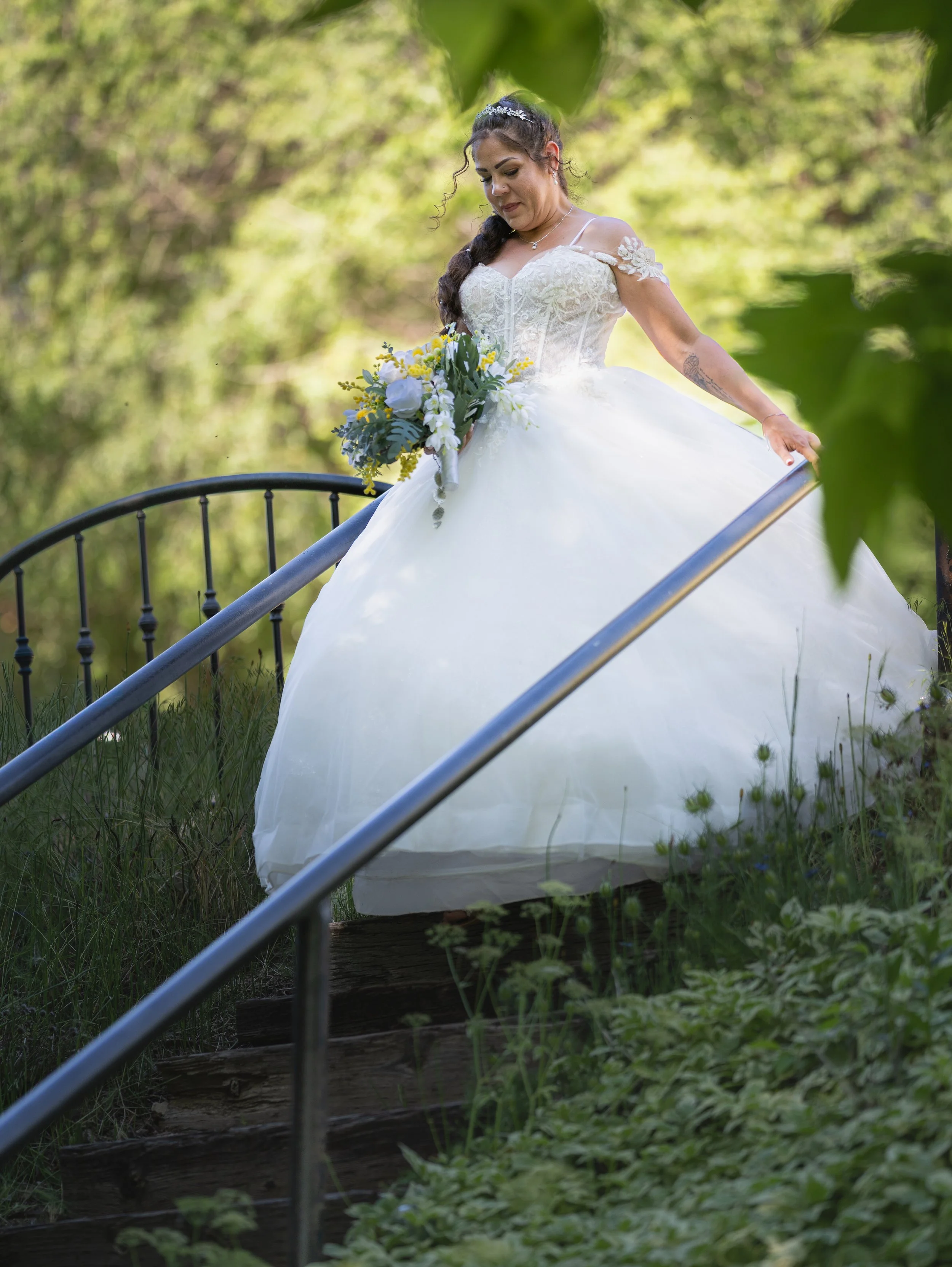 A bride in a white wedding dress holding a bouquet of flowers on a garden staircase surrounded by greenery.