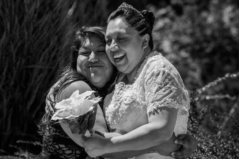 Two women embracing and smiling outdoors, one holding a large flower.