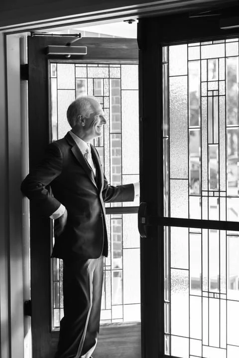 A man in a suit standing by a glass door, looking outside with a smile.