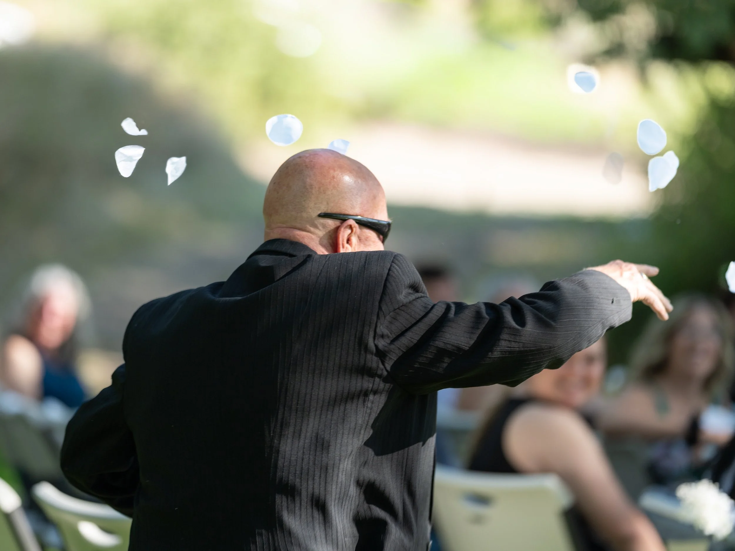Bald man in black suit with glasses at an outdoor event, raising his right arm, with blurred people and green foliage in the background.
