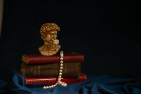 A bust of a person, stacked books, and a pearl necklace on dark fabric background.