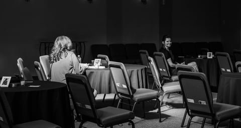 Two women sitting at separate tables in a conference room with empty chairs around them