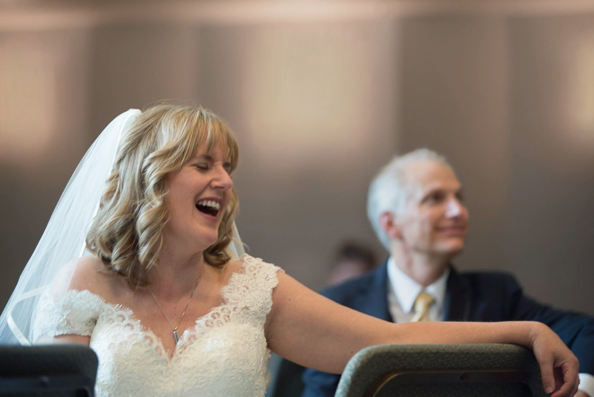 A woman in a white wedding dress with a veil, smiling and laughing, seated next to a man in a suit with a yellow tie, in a formal indoor setting.