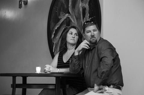 A man and a woman sitting at a tall table in a cafe or restaurant, with a candle and wine glass on the table, and a large artwork or mirror on the wall behind them.