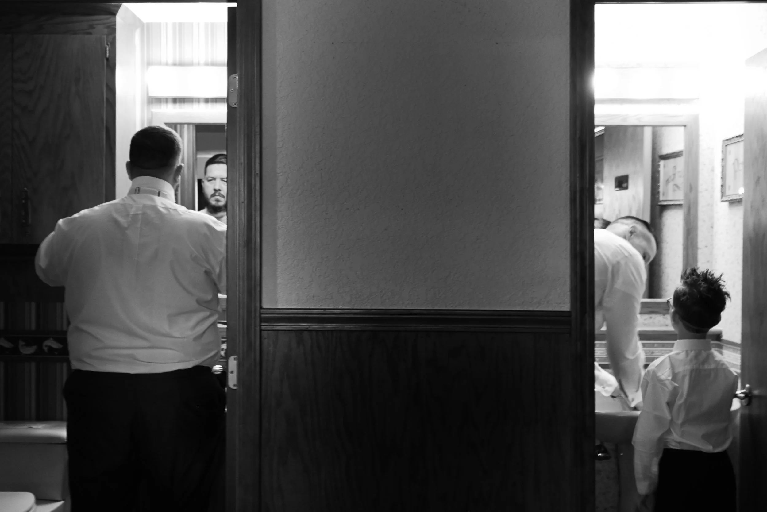 A black-and-white photo of a man in a white shirt looking into a mirror, with only the back of his head visible. Inside the mirror's reflection, a bearded man with glasses looking at the camera, and a woman with long hair can be seen. The scene appea