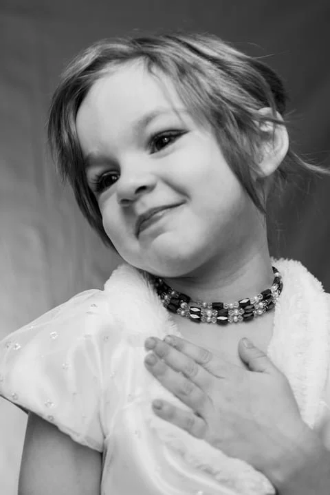 A young girl with short hair, wearing a beaded necklace and a satin dress, smiling softly with her hand on her chest in a black-and-white photograph.