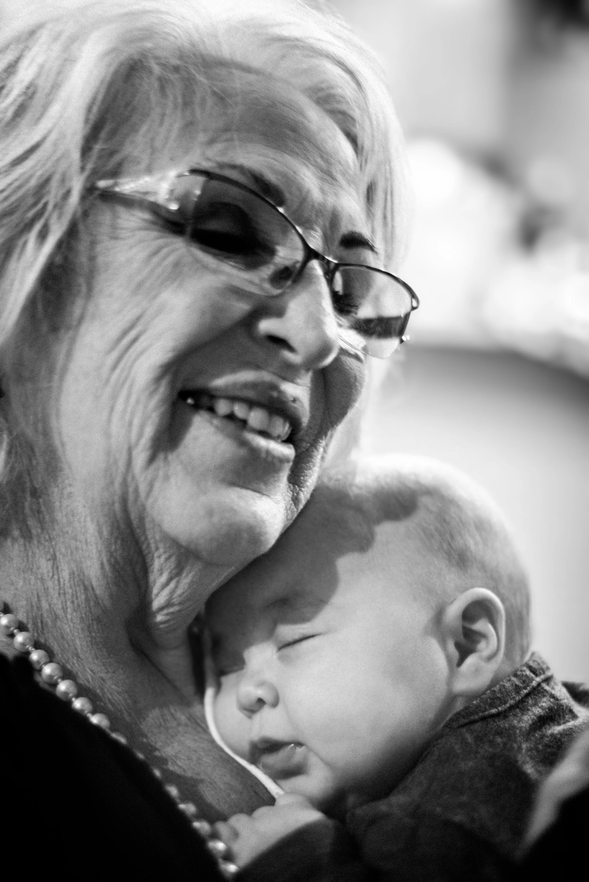 A smiling elderly woman wearing glasses, holding and cuddling a sleeping baby with a nose tube, both appearing happy in a close-up black and white photo.