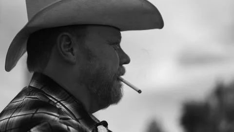 A man wearing a cowboy hat and plaid shirt smoking a cigarette in profile.