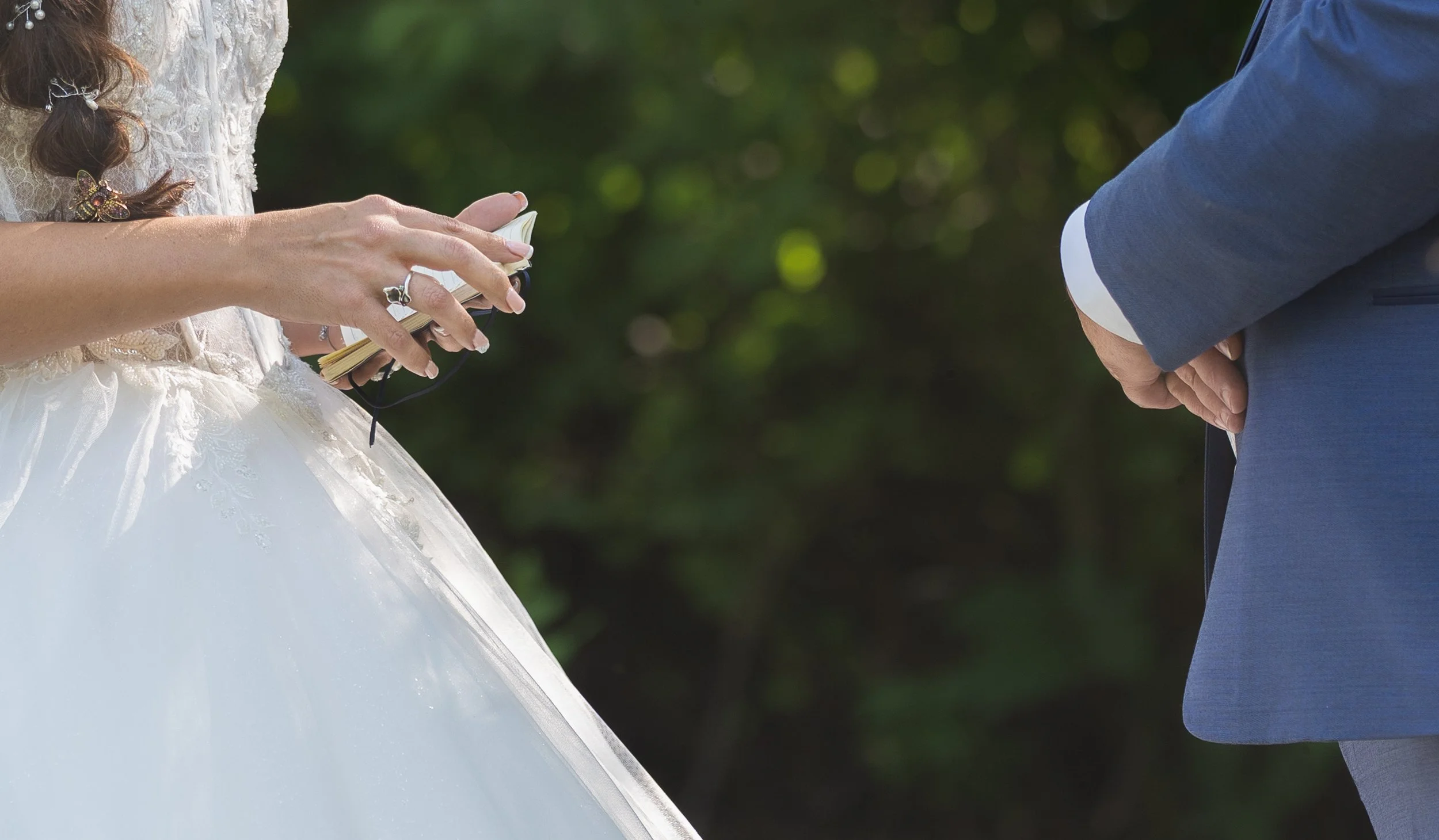 A wedding ceremony where a bride, dressed in a white lace wedding gown, is holding a book or notes and is standing next to a groom dressed in a blue suit, with their hands clasped in front of them. The background features green trees.