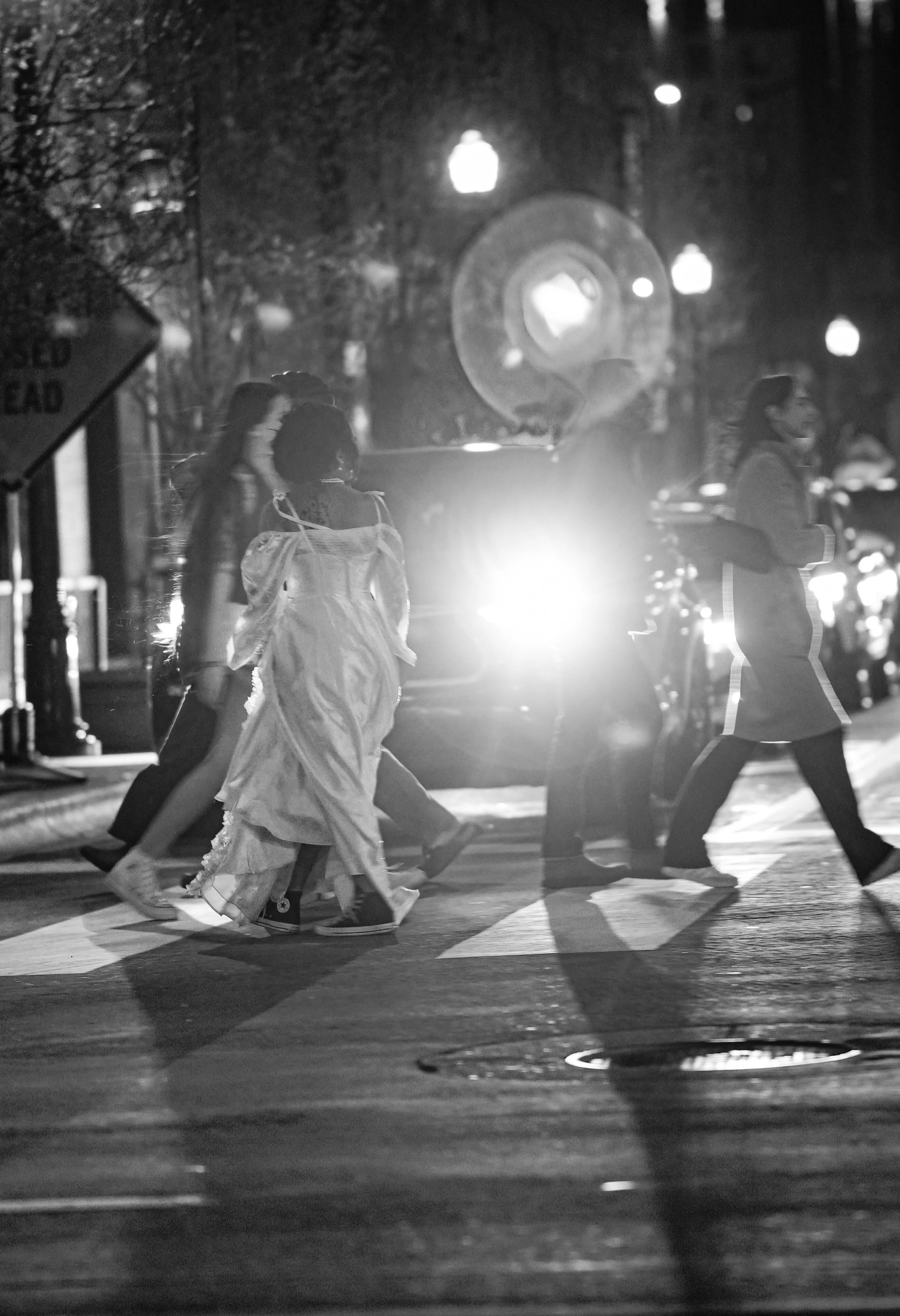 Black and white photo of people crossing a city street at night, illuminated by streetlights and car headlights.