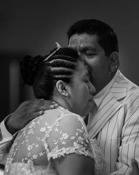A man in a striped suit hugs a woman with braided hair and a floral dress, both with eyes closed, sharing an emotional moment.