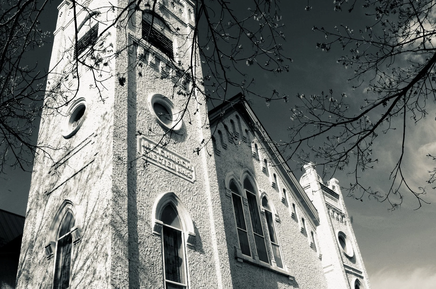 Old white building with arched windows, a sign reading 'SCHOOL NEAR 1883,' and leafless tree branches in the foreground, captured in black and white.