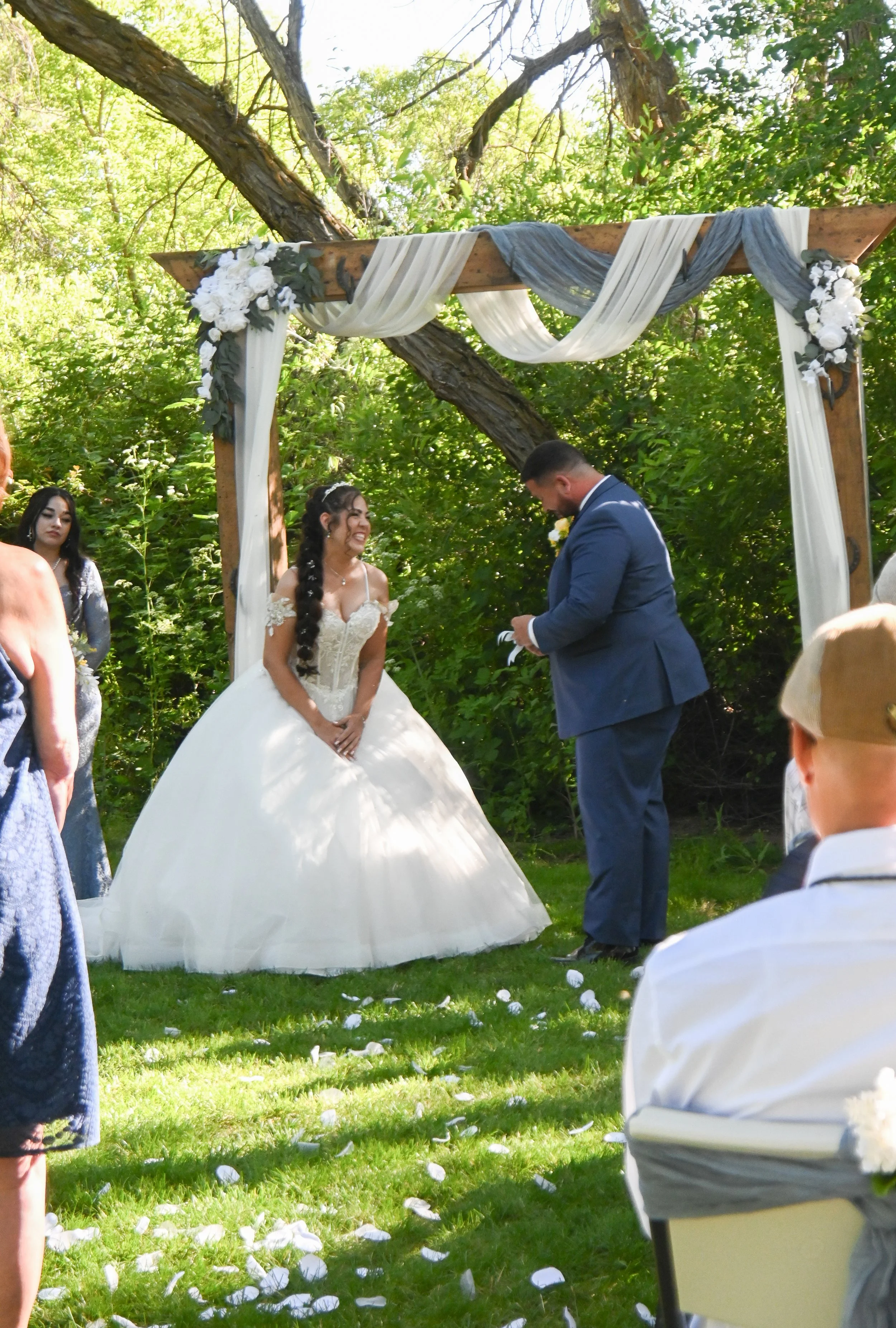 A wedding ceremony taking place outdoors under a wooden arch decorated with white and gray drapery and white flowers, with the bride and groom exchanging vows in front of seated guests surrounded by green trees.