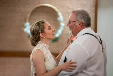 A bride and an older man, possibly her father, sharing a dance at her wedding reception, with a floral wreath hanging on the wall behind them.