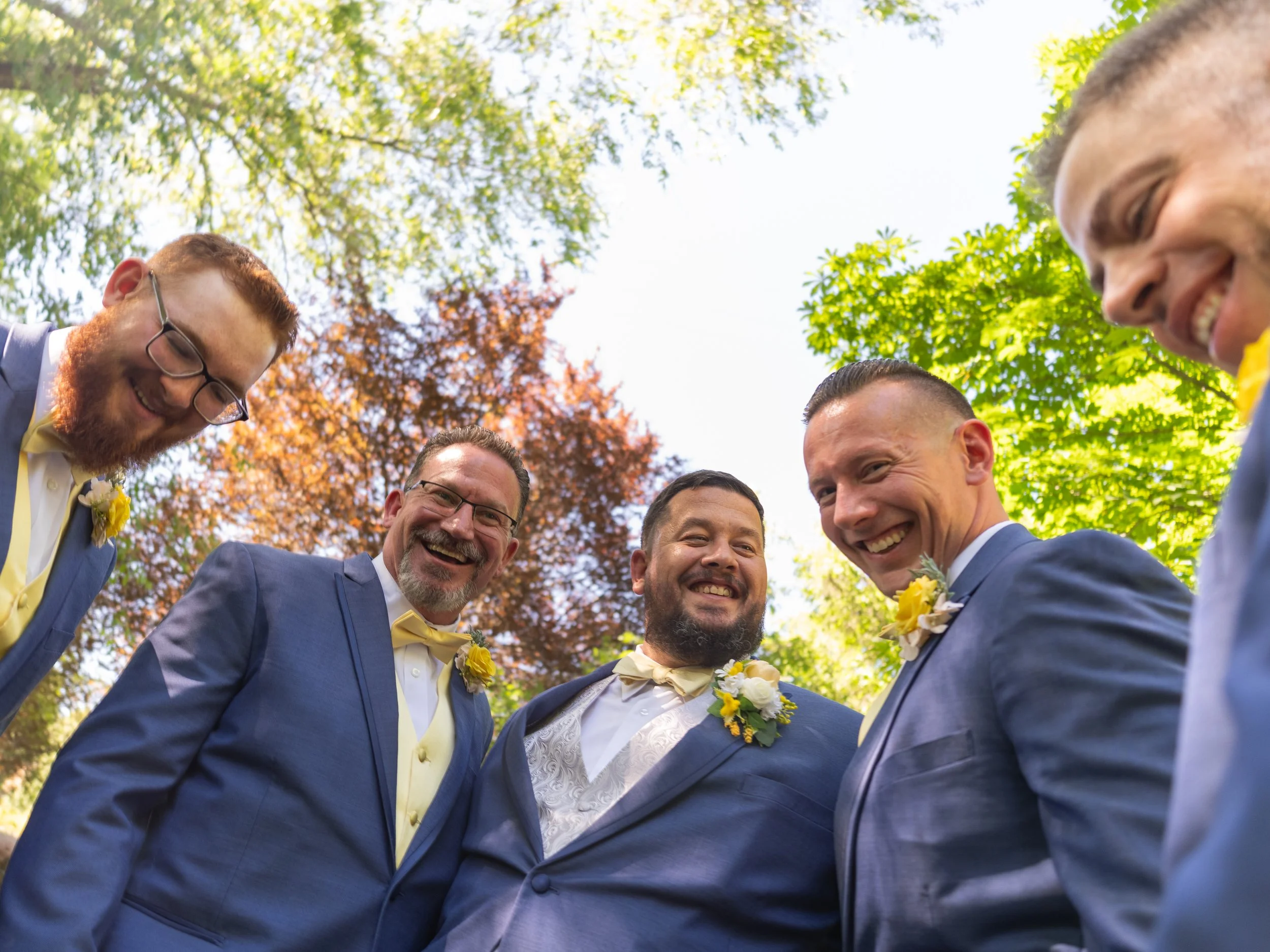 A group of men in formal suits, smiling and huddling together outdoors, with greenery and trees with autumn leaves in the background.