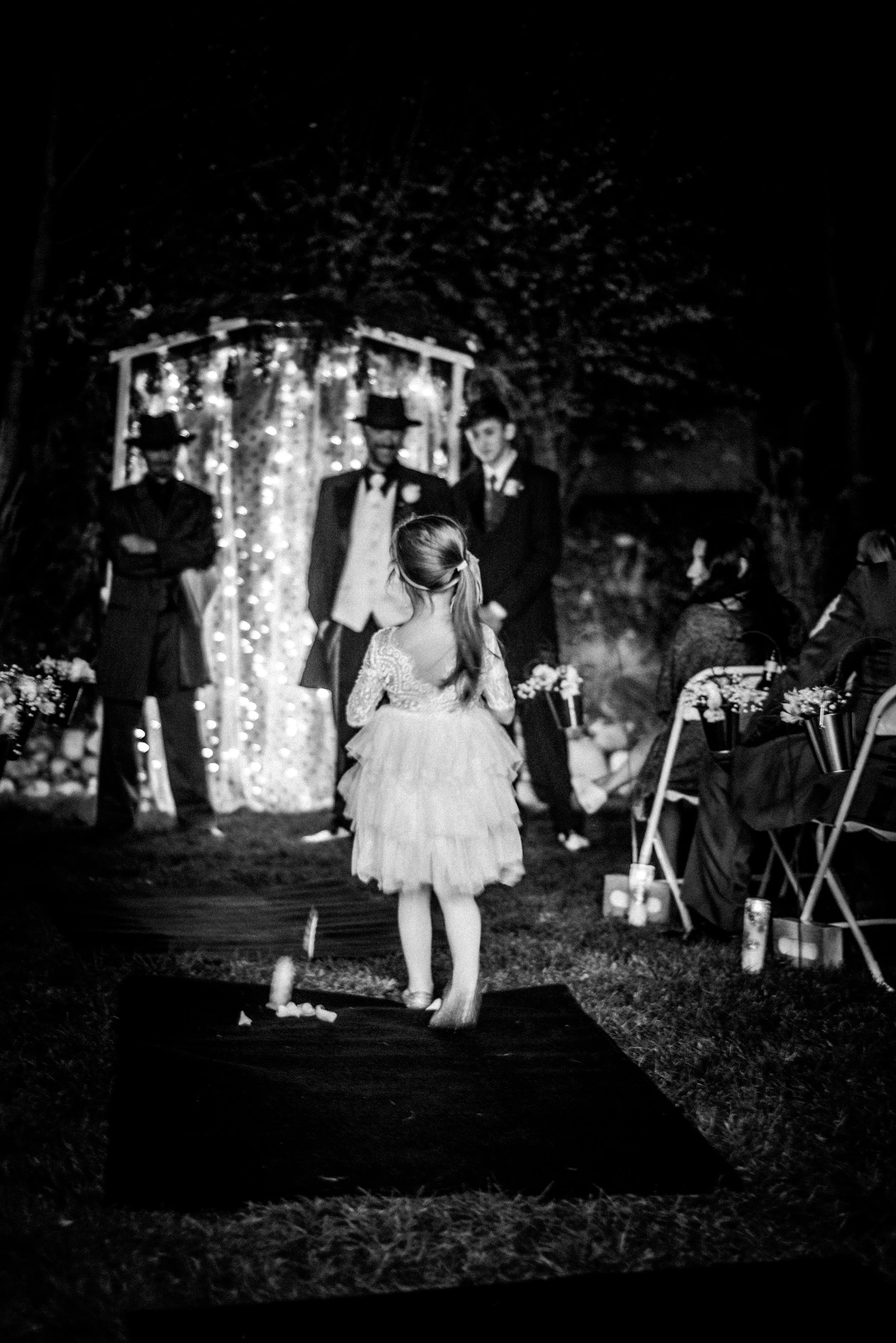 A young girl in a dress standing on a platform in front of a wedding ceremony, with three men in suits and hats, and a seated audience, at night under string lights.