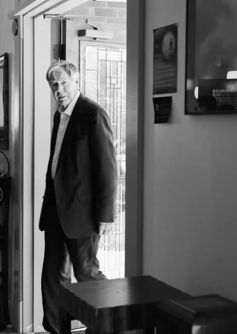 A man in a suit standing in a doorway, looking towards the camera, inside a room with framed pictures on the wall.