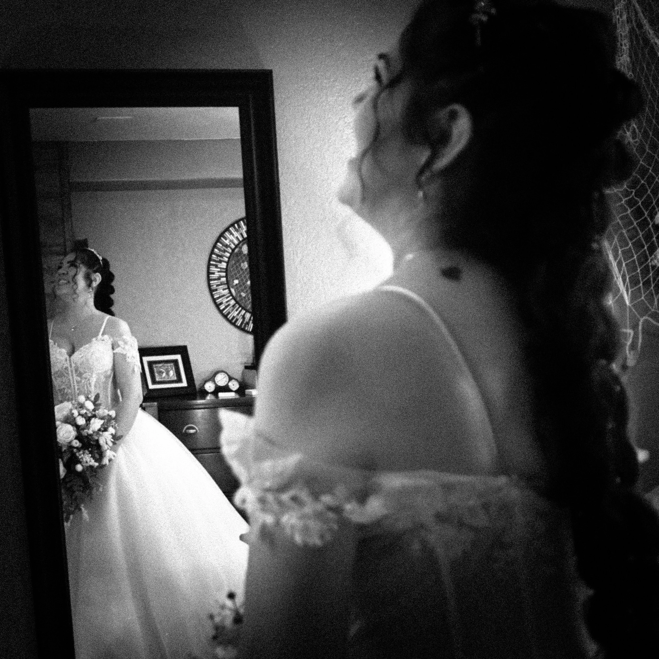 A young woman with glasses and dark hair styled in a braid, looking at a mirror, wearing a patterned shirt, in a dimly lit room with a wall clock and framed pictures in the background.