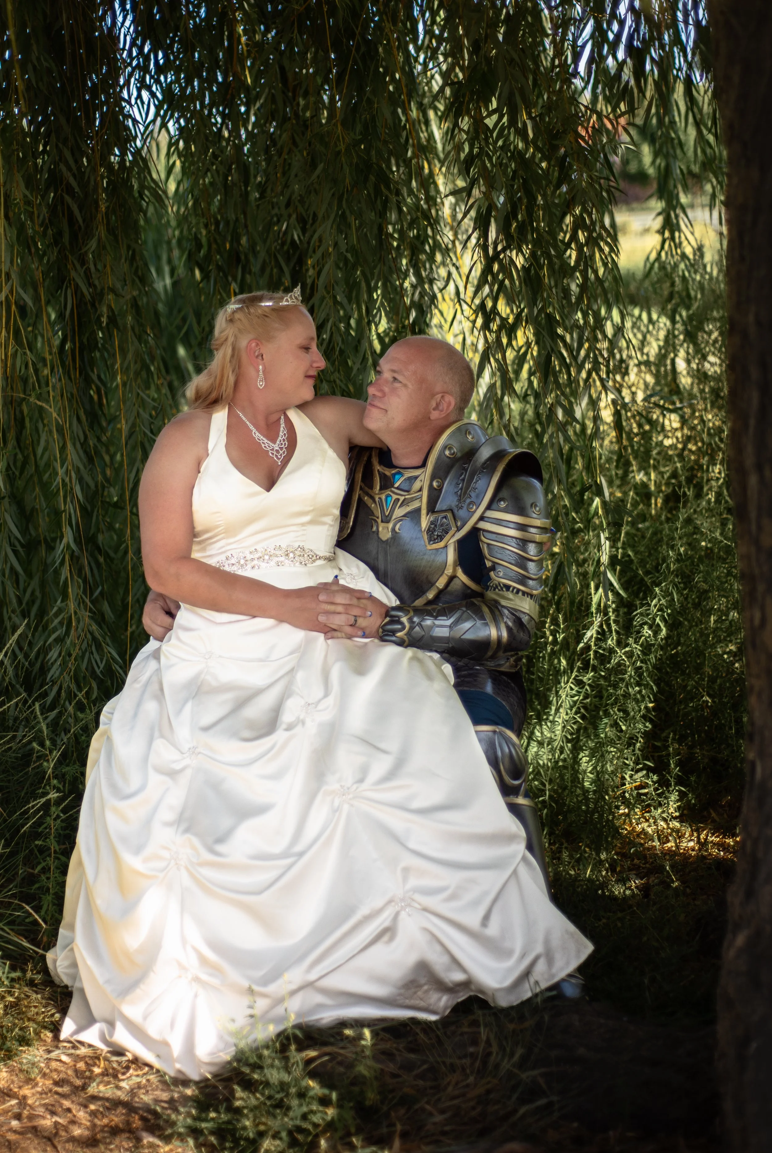 A bride in a white wedding dress and a groom in knight armor sitting among greenery, looking into each other's eyes in an outdoor setting.