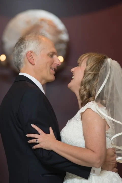 A bride and groom share a close moment at their wedding, smiling at each other, indoors with soft lighting.