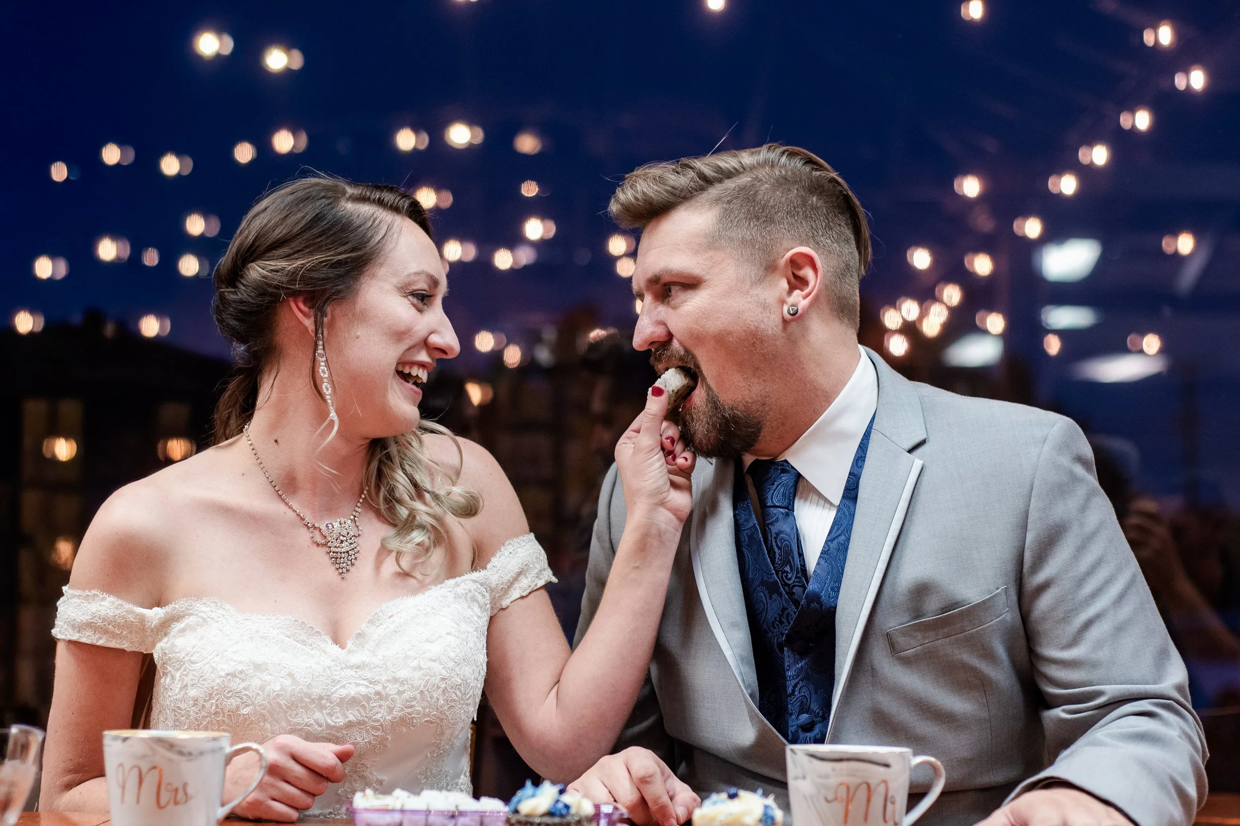Bride and groom laughing at a wedding reception, with the bride feeding a piece of cake to the groom. They are dressed in wedding attire, with string lights in the background.