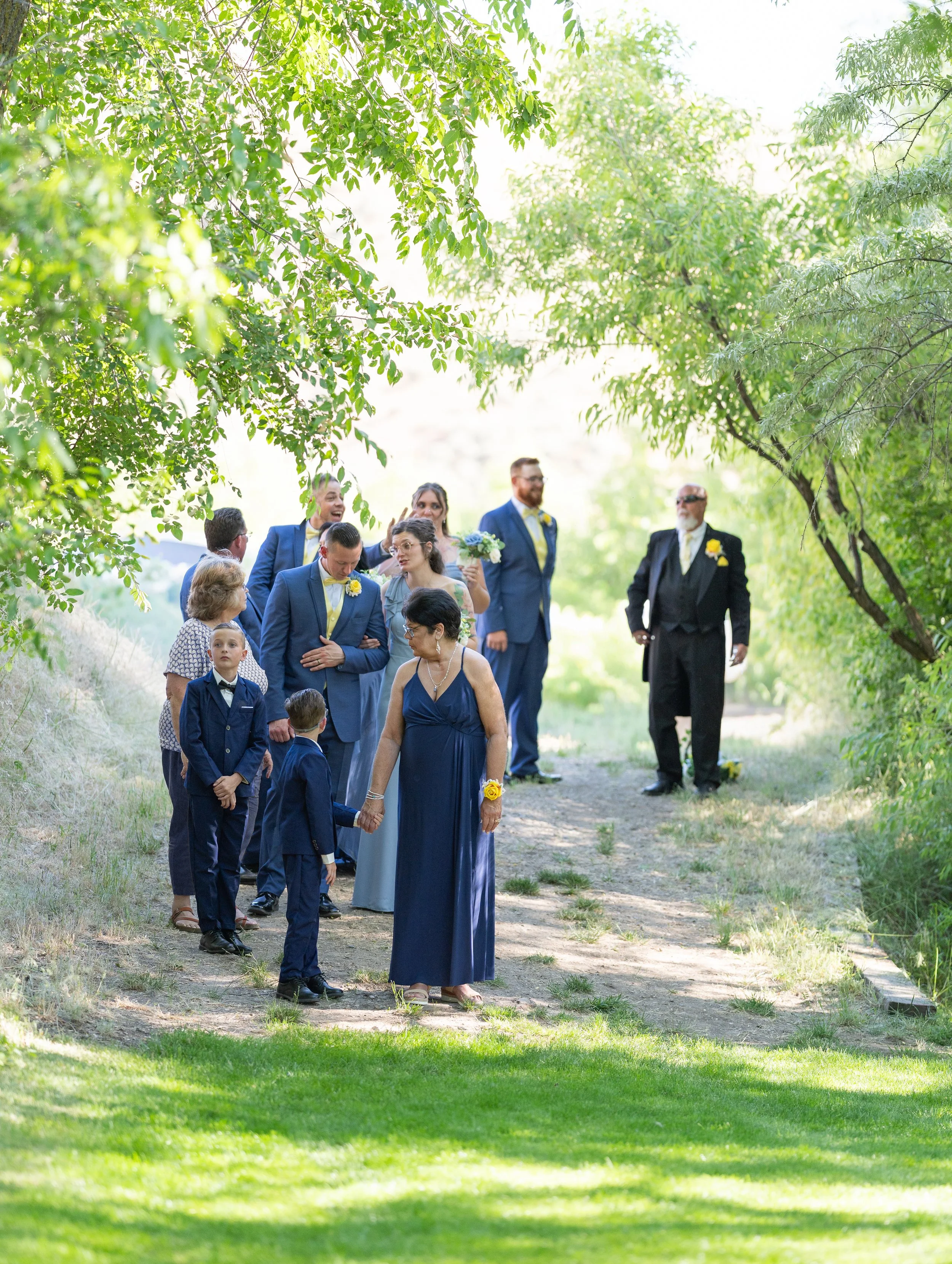 Wedding party gathers outdoors under green trees, with women in blue dresses and men in suits, some with yellow flowers, during daytime.