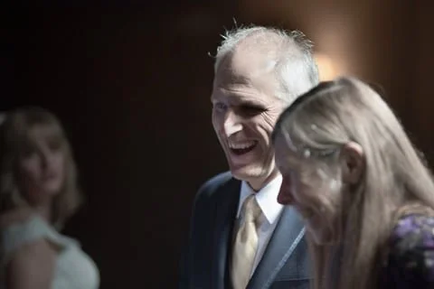 An elderly man and woman smiling and laughing together, with a woman in the background.