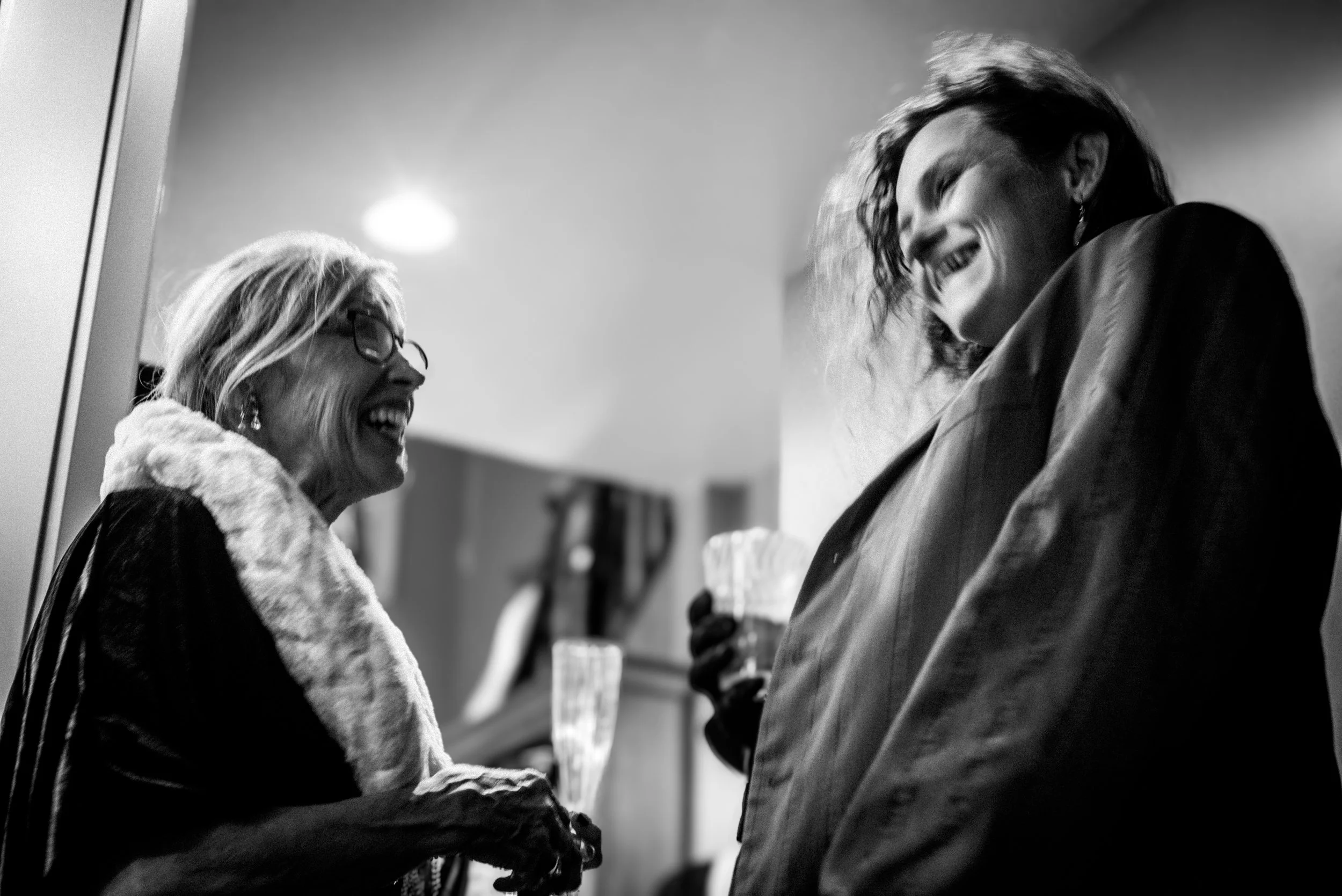 Two women are smiling and talking at an indoor social gathering, one wearing glasses and a fur collar, and the other with curly hair in a blazer, holding drinks.