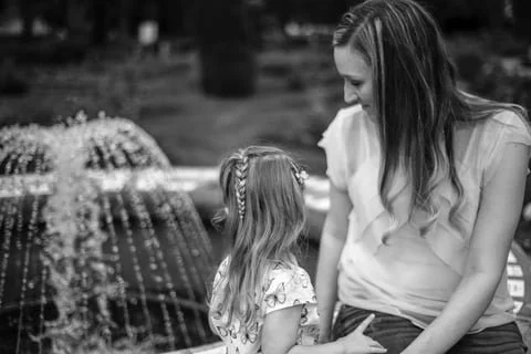 A woman and a young girl with long hair standing near a fountain in a park, smiling at each other.