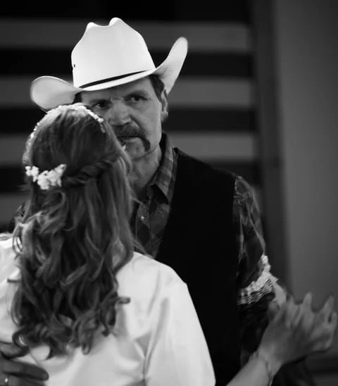 A black and white photo of a man wearing a cowboy hat and plaid shirt, dancing with a woman with long wavy hair and a floral hair accessory.