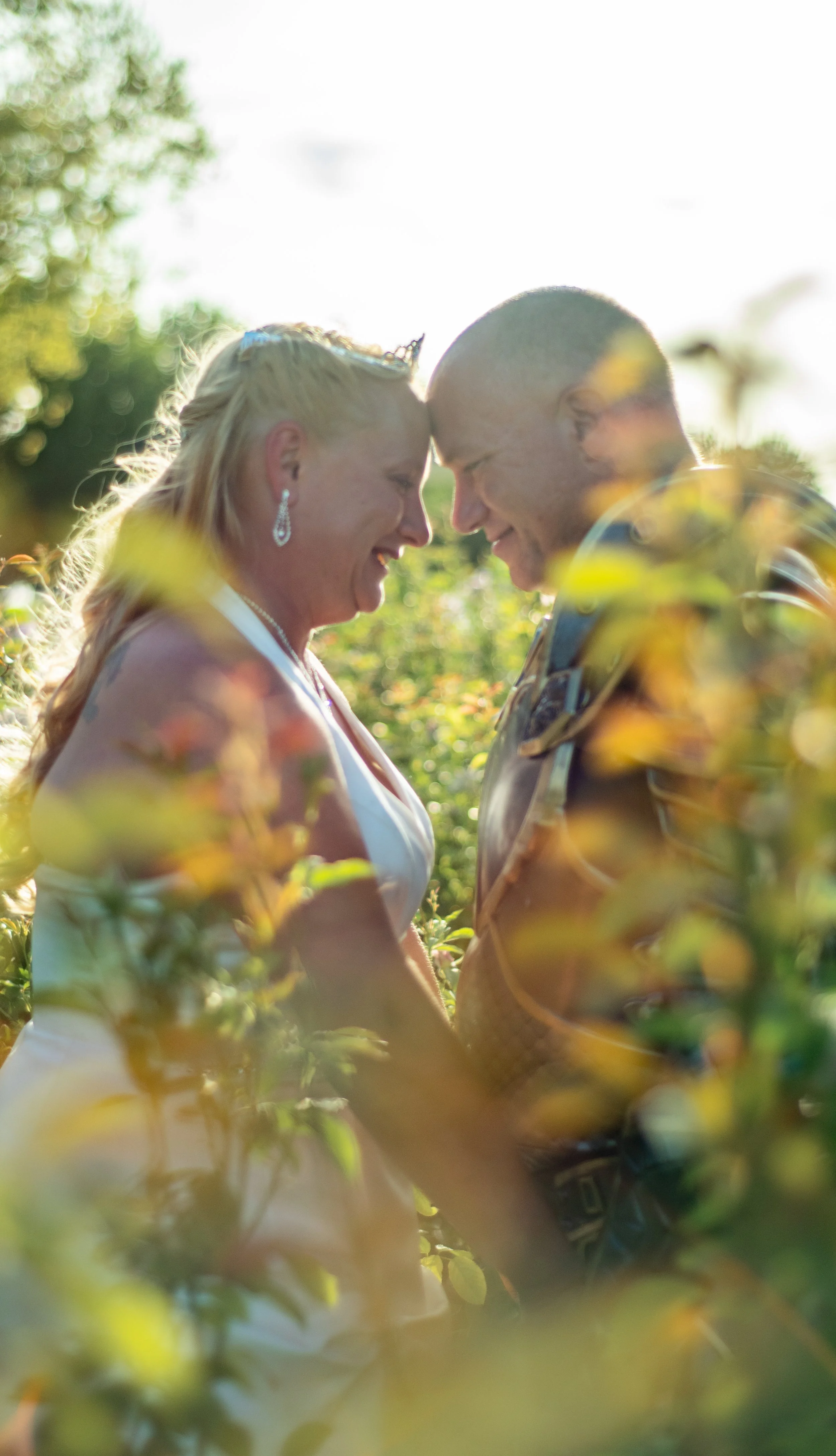 A couple standing closely, with foreheads touching, smiling and holding hands in an outdoor setting surrounded by greenery, illuminated by sunlight.