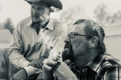 Two men, one wearing a cowboy hat and the other with glasses, sitting outdoors.