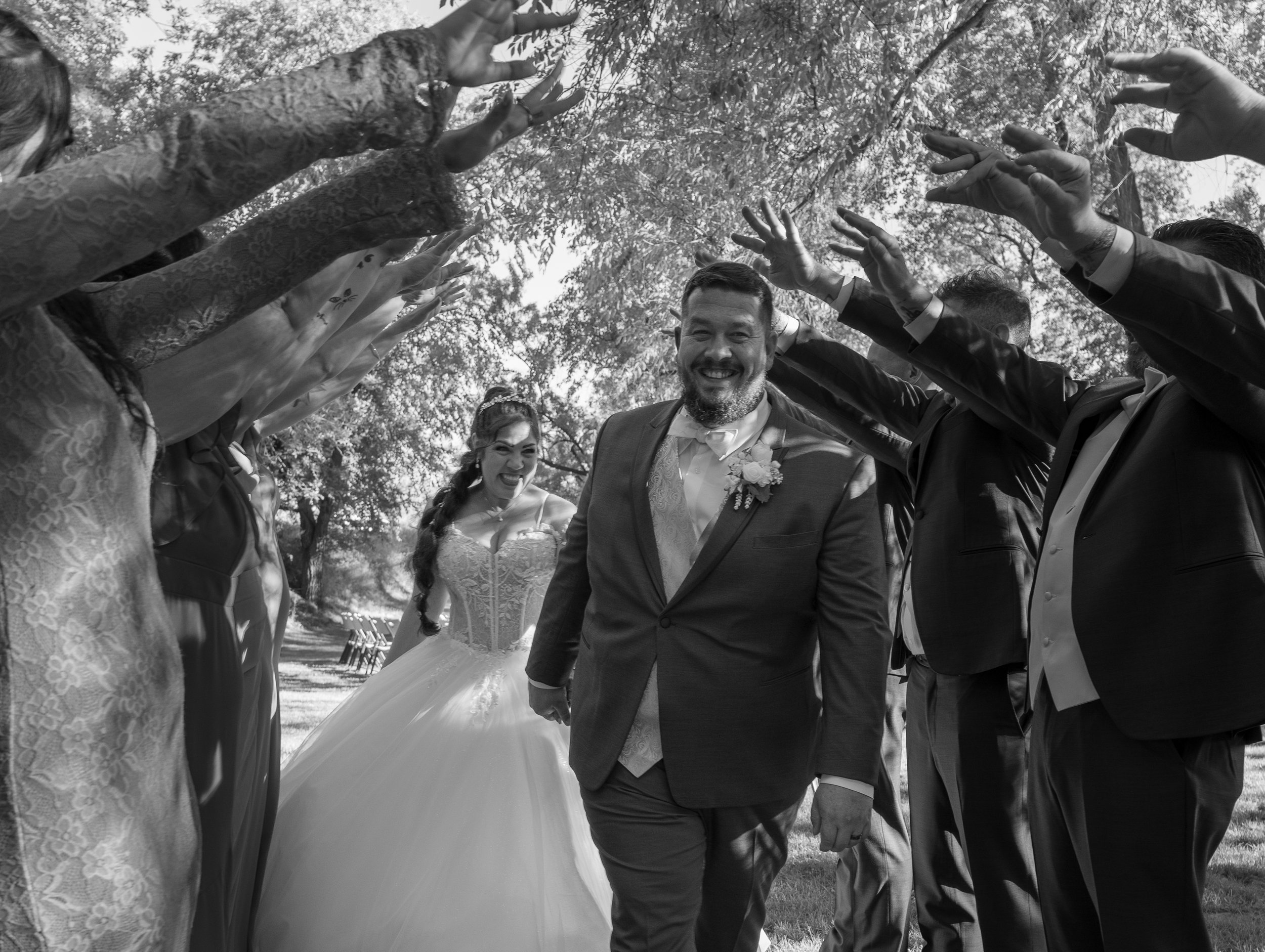 A black-and-white photo of a bride and groom at their wedding, smiling and holding hands in an outdoor setting with trees and chairs in the background. Guests surround them with their hands raised, celebrating.