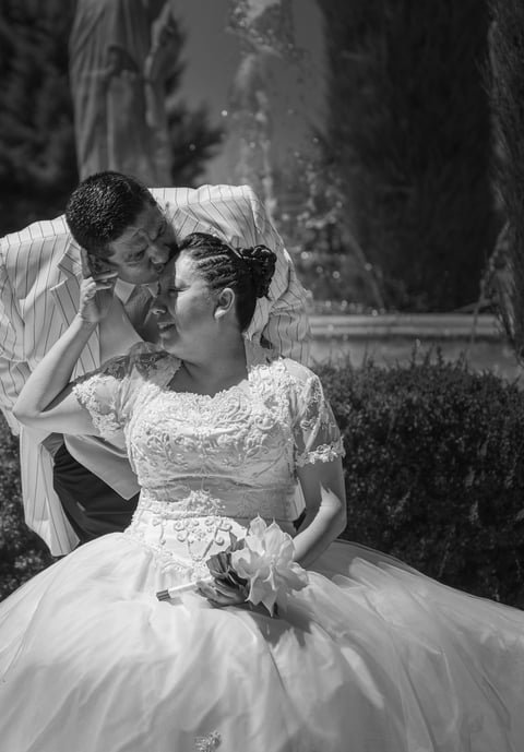 A black and white photo of a couple, with the woman sitting in a wedding dress holding a bouquet and the man leaning in close, sharing an intimate moment outdoors.