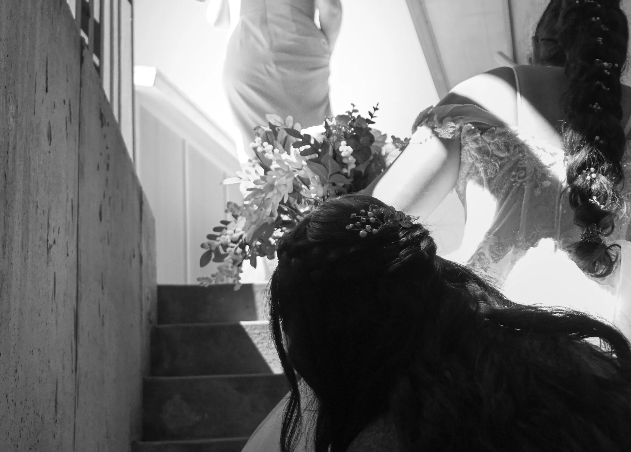 A black-and-white photo of a woman with long braided hair, wearing a lace dress, holding a bouquet of flowers.