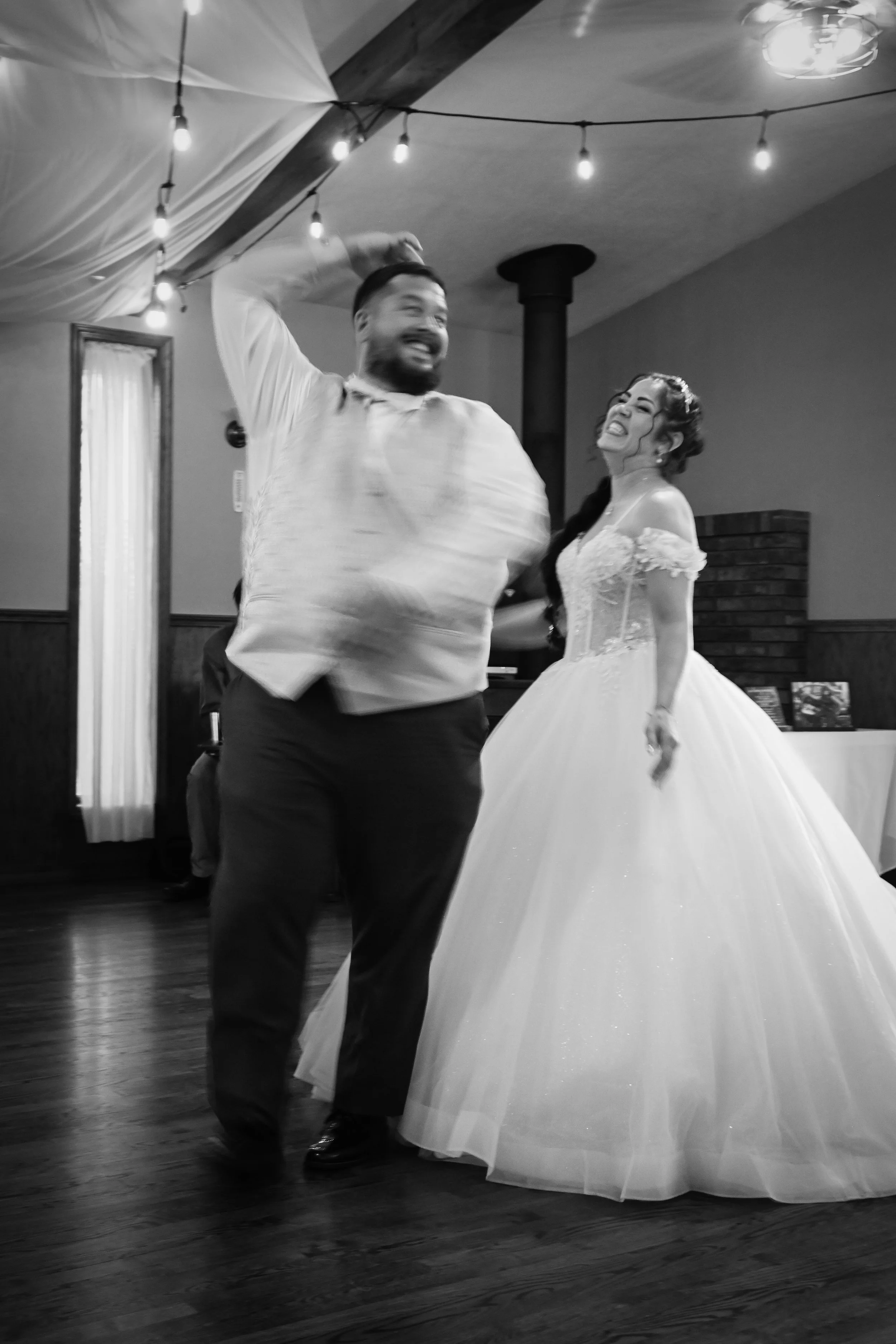 A man and woman are dancing at a wedding reception. The man is wearing a light-colored shirt and dark pants, and the woman is in a wedding gown. They are smiling and holding hands, with string lights hanging from the ceiling in the background.