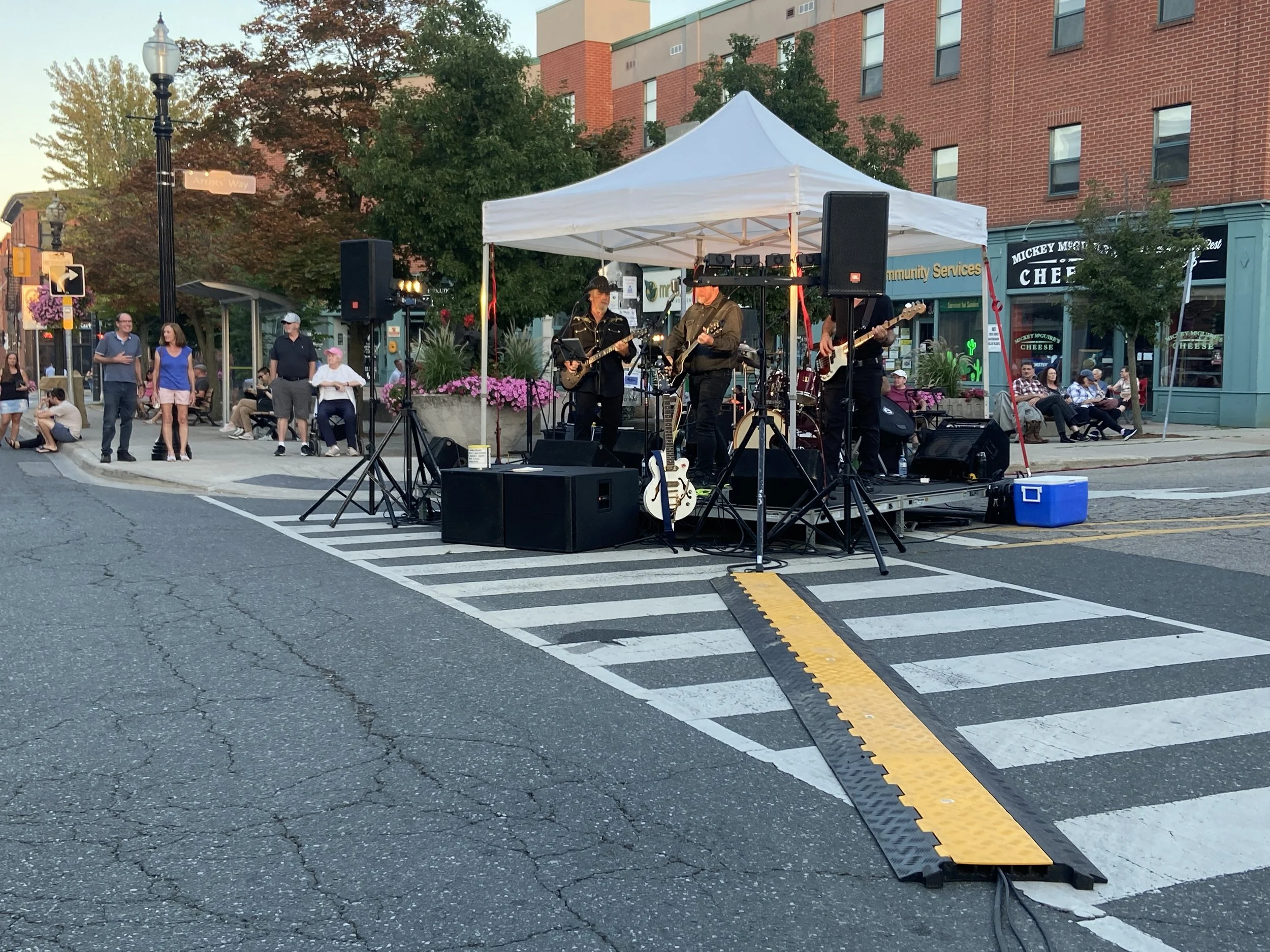 A small outdoor stage set up on a city street with a band performing under a white canopy. The band has at least three members with guitars and a drum set behind them. Several people are watching the performance, sitting on benches or standing nearby