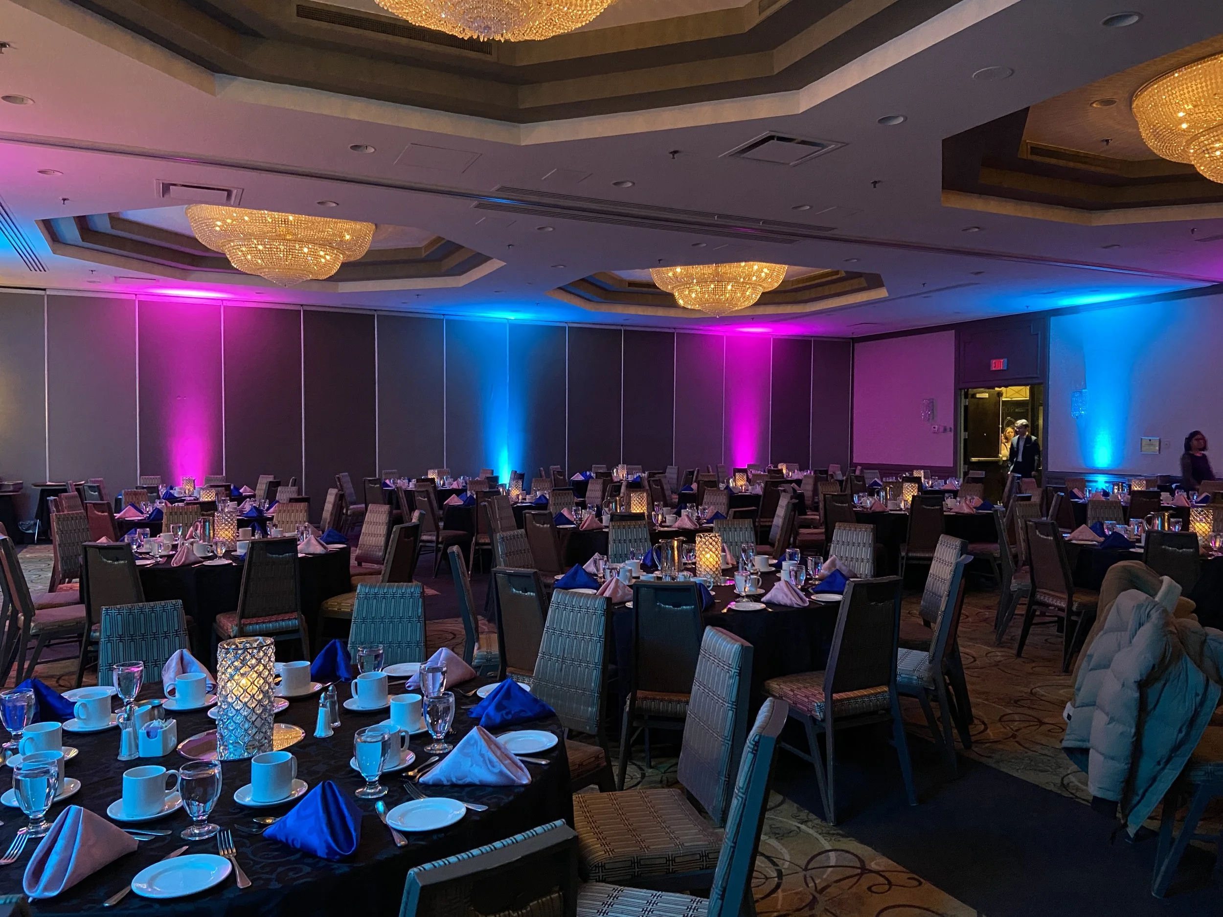 A large banquet hall decorated with colorful lighting on the walls, set with round tables covered in black tablecloths, arranged with plates, cups, glasses, napkins, and lit candle centerpieces, ready for an event.