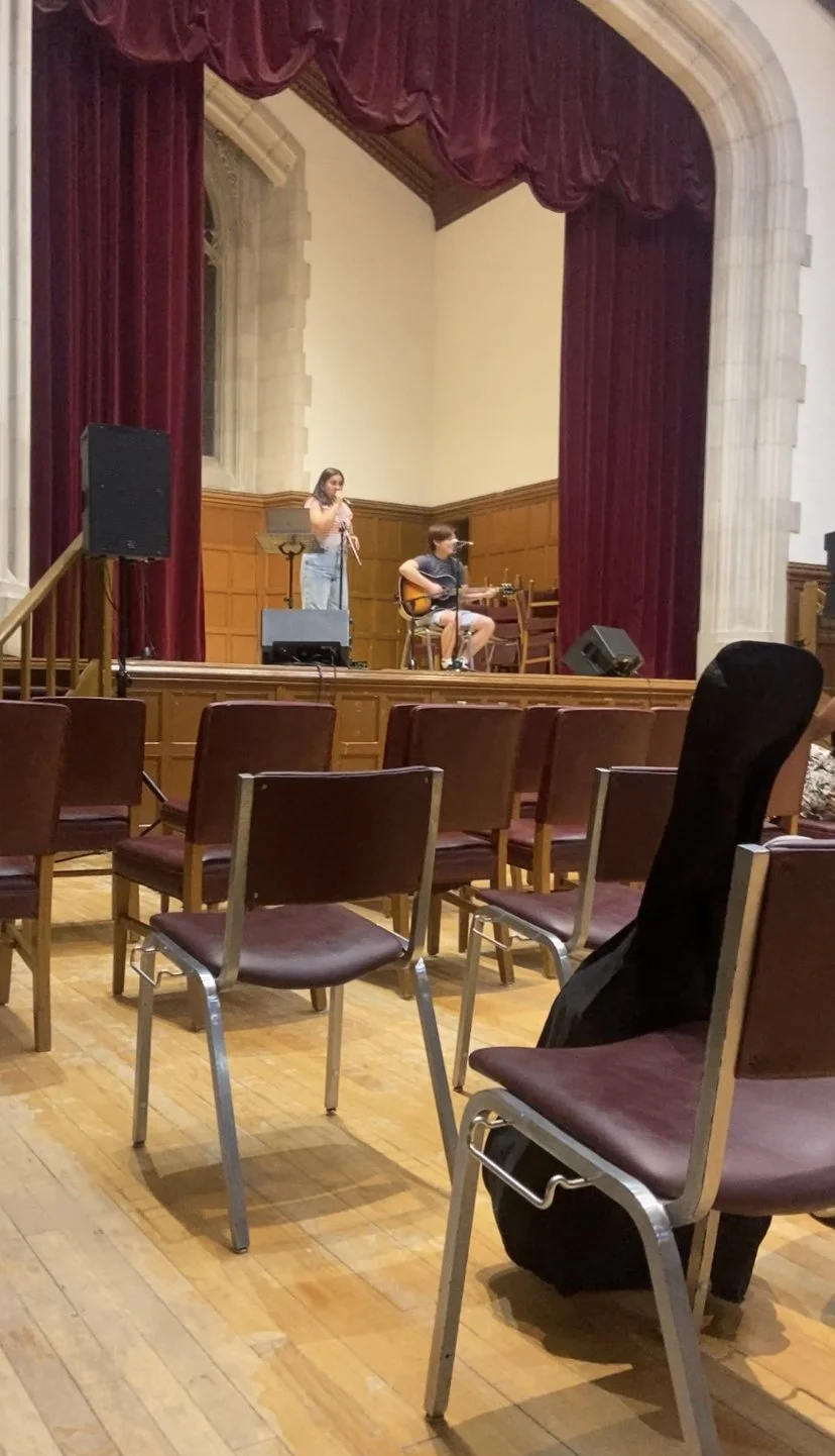 A performance on stage inside a theater with two musicians, one playing guitar and another singing, with empty chairs in the foreground.